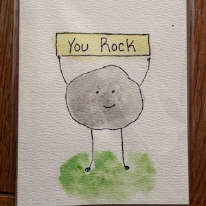 May include: A watercolor greeting card featuring a smiling gray rock character holding a yellow sign that reads "You Rock." The rock stands on a patch of green grass, all set against a white background.