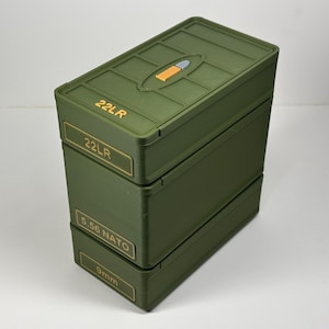 May include: Stacked olive green storage boxes with labels for ammunition types. The top box has a small orange and blue design. Labels read "22LR", "5.56 NATO", and "9mm".