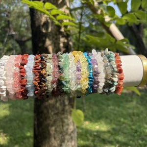 Può includere: Una collezione di braccialetti con schegge di pietre preziose in vari colori, tra cui bianco, rosa, rosso, marrone, blu, verde, giallo, viola e arancione. I braccialetti sono esposti su un cilindro bianco con una fascia dorata, mettendo in risalto le texture delle pietre naturali.
