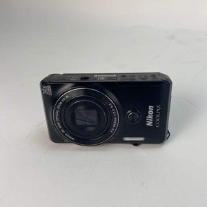 May include: A black Nikon COOLPIX digital camera with a zoom lens. The camera has a rectangular shape with rounded corners. The lens is centered on the front, with the Nikon COOLPIX logo on the right side. The camera has a 12x wide optical zoom.