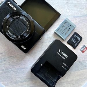 May include: Black Canon G7X digital camera with a zoom lens, open screen, and accessories including a battery, charger, and SD card. The charger indicates 'CHARGE' and 'FULL'.