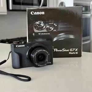 May include: A black Canon PowerShot G7 X Mark III digital camera with a zoom lens, and its box. The box shows the camera's features and the Canon logo. The camera has a black strap.