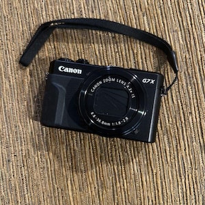 May include: A black Canon G7 X digital camera with a zoom lens and a black wrist strap. The camera has the Canon logo in white. The lens has the text "CANON ZOOM LENS 4.2x IS" and "8.8-36.8mm 1:1.8-2.8".