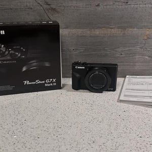 May include: A black Canon PowerShot G7 X Mark III digital camera with its box and paperwork. The camera has a lens and control dials. The box is black with the Canon logo and camera image. The camera is on a white surface.