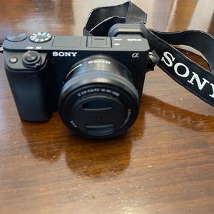 May include: Black Sony Alpha digital camera with a lens and a black strap with the word "SONY" in white. The camera is on a wooden surface. The lens has a black lens cap.