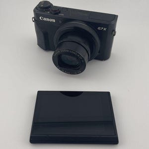 May include: A black Canon G7 X digital camera with a zoom lens and the Canon logo. Below the camera is a black rectangular case. The camera has a lens that reads "CANON ZOOM LENS 4.2x IS".