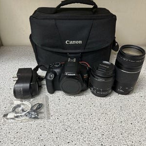 May include: A black Canon EOS camera kit including a bag, two lenses, a charger, and cables. The camera and lenses are black with Canon branding. The bag is black with a handle and the Canon logo.