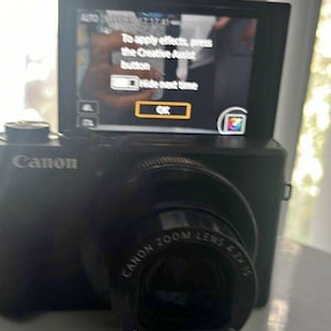 May include: Black Canon digital camera with a zoom lens and a screen displaying a menu. The screen shows the text "To apply effects, press the Creative Assist button" and "OK".