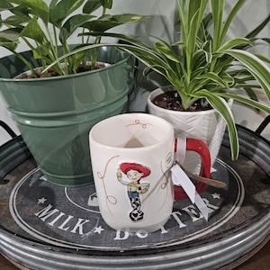 May include: A white ceramic mug featuring a raised graphic of the character Jessie from Toy Story, with a red handle and a small white tag attached. The mug is decorated with thin red swirling lines and a small red swirl near the inner rim.