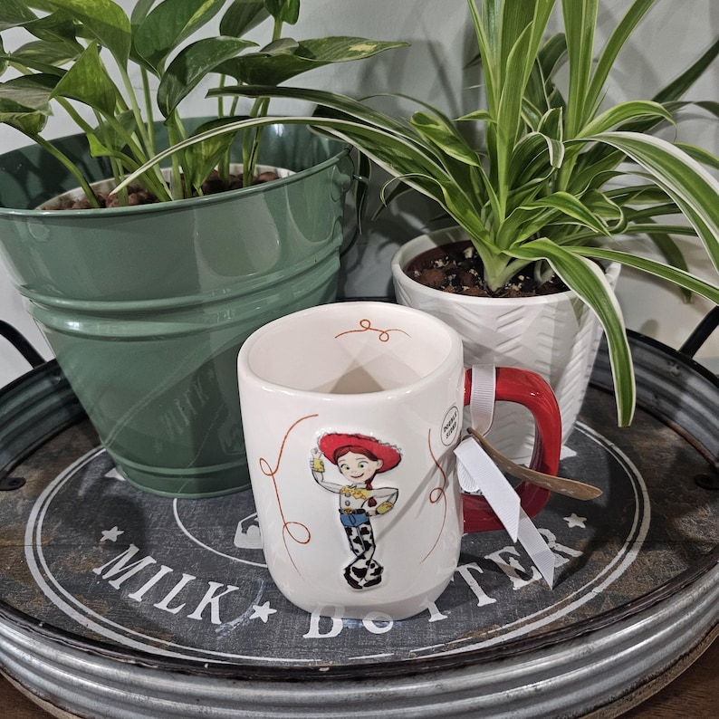 May include: A white ceramic mug featuring a raised graphic of the character Jessie from Toy Story, with a red handle and a small white tag attached. The mug is decorated with thin red swirling lines and a small red swirl near the inner rim.