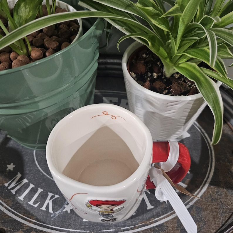May include: A white ceramic mug featuring a red handle and a graphic of a character with a red hat. The interior rim has a small orange swirl design. A white ribbon is tied to the handle. The mug is positioned in front of two potted plants.