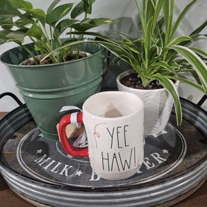 May include: A white ceramic mug with a bright red handle and the text YEE HAW! printed in black lettering. The mug features small red decorative swirls on the sides and a white ribbon tied around the handle, positioned on a round metal tray.