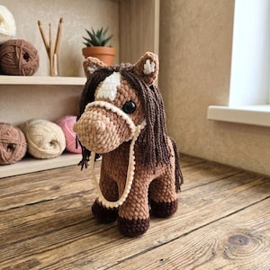 Puede incluir: Un juguete de caballo de ganchillo hecho a mano en tonos marrones, beige y blanco. El caballo tiene una mancha blanca en la frente, una melena marrón oscuro y un cabestro blanco. El juguete está sobre una superficie de madera.