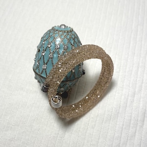 May include: A light brown crystal bracelet lies beside a turquoise egg-shaped jewellery box. The bracelet is made of small, sparkling crystals. The jewellery box has silver-coloured trim and is decorated with clear crystals.