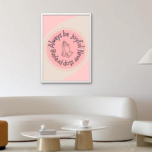Puede incluir: Una impresión artística enmarcada con un fondo rosa y beige. La impresión presenta un diseño circular con manos rezando y el texto "Always be joyful, never stop praying". La obra de arte se muestra en un marco blanco.