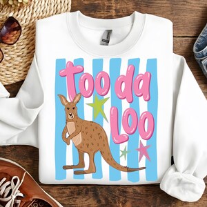 Może przedstawiać: Biała bluza z ilustracją kangura i napisem "Tooda Loo" w kolorze r&oacute;żowym. Kangur jest brązowy z jasnym brzuchem, na tle niebieskich pask&oacute;w i zielonych i r&oacute;żowych gwiazd. Zabawny, swobodny design.