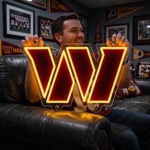 May include: A Washington Commanders logo neon sign featuring a large letter W in deep burgundy with a bright yellow glowing outline. The sign is constructed from clear acrylic and includes circular mounting holes at the top and bottom edges.