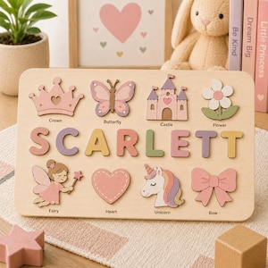 May include: A rectangular wooden name puzzle featuring the name SCARLETT in large, pastel-colored letters. The board includes cutouts for a crown, butterfly, castle, flower, fairy, heart, unicorn, and bow, each labeled with its corresponding name.