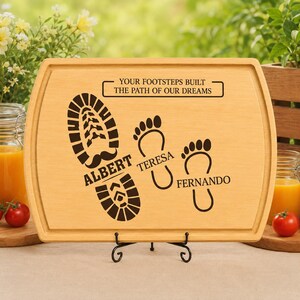 May include: A rectangular light-colored wooden cutting board featuring a dark engraved design of one large boot print labeled Albert and two smaller bare footprints labeled Teresa and Fernando, with the text YOUR FOOTSTEPS BUILT THE PATH OF OUR DREAMS.