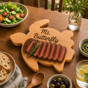 May include: A wooden turtle-shaped cutting board with the text "Ms. Butterfly" engraved on it. Sliced steak is displayed on the board, garnished with rosemary. The table setting includes a salad, bread, olives, and a glass of water.