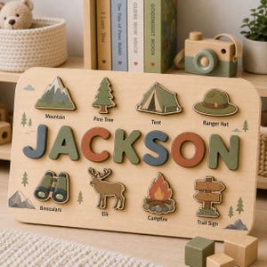 May include: A wooden name puzzle featuring the name JACKSON in large, multi-colored letters. Below and above the name are eight wooden cutout shapes including a mountain, pine tree, tent, ranger hat, binoculars, elk, campfire, and trail sign.
