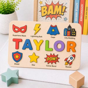 May include: Wooden puzzle featuring the name "TAYLOR" in colorful letters, with superhero-themed images like a mask, cape, and rocket. The puzzle includes the word "POW!" and is designed for educational play.