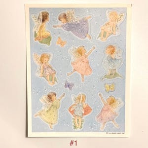 May include: A sheet of stickers featuring illustrations of fairies with glittery wings and dresses in pastel colors. Butterflies and sparkling accents are scattered across the light blue background. The number "#1" is printed at the bottom.