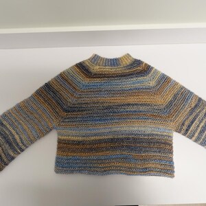 Op de afbeelding: Een handgehaakte trui met een hoge halslijn en uitlopende mouwen. De trui heeft horizontale strepen in tinten blauw, bruin en bruin. Het kledingstuk wordt gepresenteerd op een wit oppervlak.