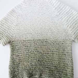 Op de afbeelding: Een handgehaakt shirt met korte mouwen en een kleurengradatie. Het bovenste gedeelte is licht, gebroken wit, dat overgaat in een gedempt groen aan de onderkant. Het shirt heeft een geribbelde textuur op de mouwen.