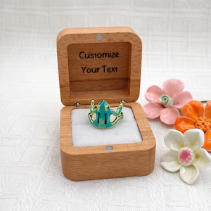 May include: A wooden ring box with the text "Customize Your Text" engraved inside. The box is open, revealing a turquoise frog ring with gold accents. Decorative ceramic flowers in pink, orange, and white are arranged nearby.