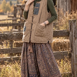 May include: A tan quilted vest with a high collar, toggle closures, and dark gray patch accents, worn over a long-sleeved olive green shirt. The outfit includes a loose-fitting, ankle-length skirt featuring a multi-colored floral pattern.