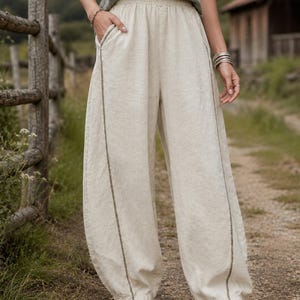 Könnte beinhalten: Cremefarbene, weit geschnittene Hose mit elastischem Bund, Seitentaschen und einer vertikalen Naht entlang der Vorderseite jedes Beins. Die Hose hat eine lockere, weit ausgestellte Silhouette, die sich zum Knöchel hin leicht verjüngt.