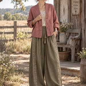 May include: A dusty rose long-sleeve button-down jacket with a V-neckline, worn over a cream-colored sleeveless top. The outfit includes olive green wide-leg trousers featuring a drawstring waist with tassels and vertical floral embroidery.