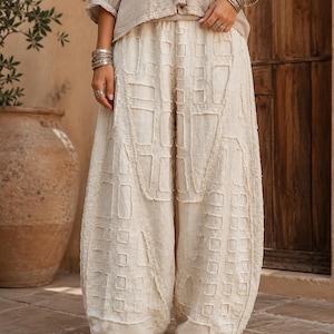 Op de afbeelding: Off-white losvallende broek met een gestructureerd patchwork-achtig ruitpatroon met gerafelde randen. De broek heeft een high-waisted, volumineus silhouet dat bij de enkels iets toeloopt, gecombineerd met een bijpassende lichtbeige top.