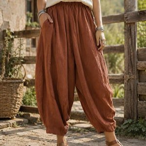 May include: Loose-fitting, rust-colored trousers featuring an elasticized waistband, a gathered silhouette, and side pockets. The pants are crafted from a textured fabric and fall to a cropped length, showcasing a relaxed, voluminous design.