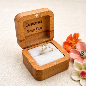 May include: A wooden ring box with a hinged lid, open to reveal a gold ring with a bunny ear design. The box is engraved with "Customize Your Text". The ring sits on a white velvet cushion. Decorative flowers are in the background.