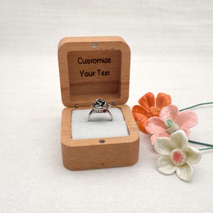 May include: A silver ring with a black rose design and red accents sits inside an open wooden ring box. The box is engraved with the words "Customize Your Text". Decorative orange, pink, and white flowers are next to the box.