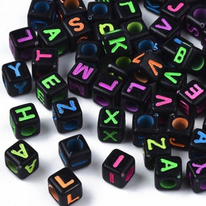 Puede incluir: Una colección de cuentas de letras negras con letras de colores neón. Las cuentas están esparcidas sobre una superficie blanca.