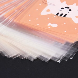 May include: A stack of clear plastic bags with a peach-coloured bag on top. The peach-coloured bag has a white cat illustration with black eyes and stripes. The cat is holding a fish.