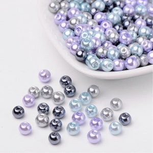 Puede incluir: Una colecci&oacute;n de cuentas de perlas en tonos lavanda, azul y gris. Las cuentas est&aacute;n esparcidas sobre una superficie blanca y en un peque&ntilde;o plato blanco.