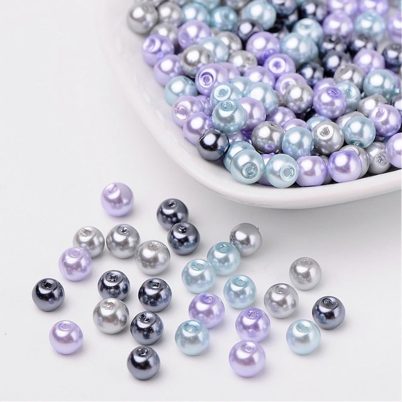 Puede incluir: Una colecci&oacute;n de cuentas de perlas en tonos lavanda, azul y gris. Las cuentas est&aacute;n esparcidas sobre una superficie blanca y en un peque&ntilde;o plato blanco.