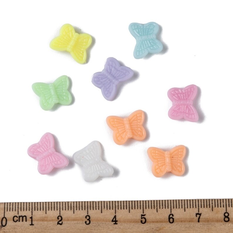 Op de afbeelding: Assortiment van tien pastelkleurige plastic vlinder kralen. De kralen zijn ongeveer 2,5 cm groot en zijn gerangschikt op een wit oppervlak met een liniaal voor schaal.