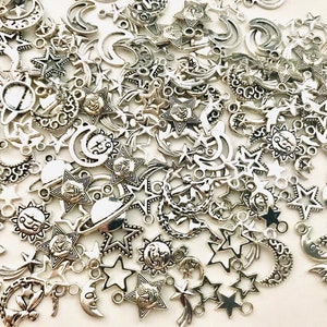 Op de afbeelding: Een grote verzameling zilverkleurige metalen bedels met sterren, manen en andere hemelmotieven. Deze bedels zijn perfect om een vleugje whimsy toe te voegen aan sieraden, knutselwerk en andere projecten.