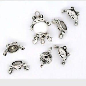 以下が含まれることがあります： 白いビーズが付いた銀色のカエルチャームのセット。チャームは座っている、立っている、跳ねているなど、さまざまなポーズをしています。チャームは、ジュエリーやクラフトに遊び心のあるタッチを加えるのに最適です。