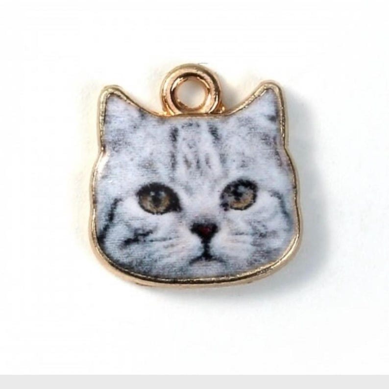 K&ouml;nnte beinhalten: Ein goldfarbener Anh&auml;nger mit einem grauen und wei&szlig;en Katzenkopf und einer goldenen &Ouml;se oben.