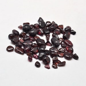 May include: A collection of polished, dark red garnet gemstones of varying sizes and shapes. The gemstones are scattered on a white surface, with some appearing translucent and others opaque. The overall appearance is smooth and reflective.