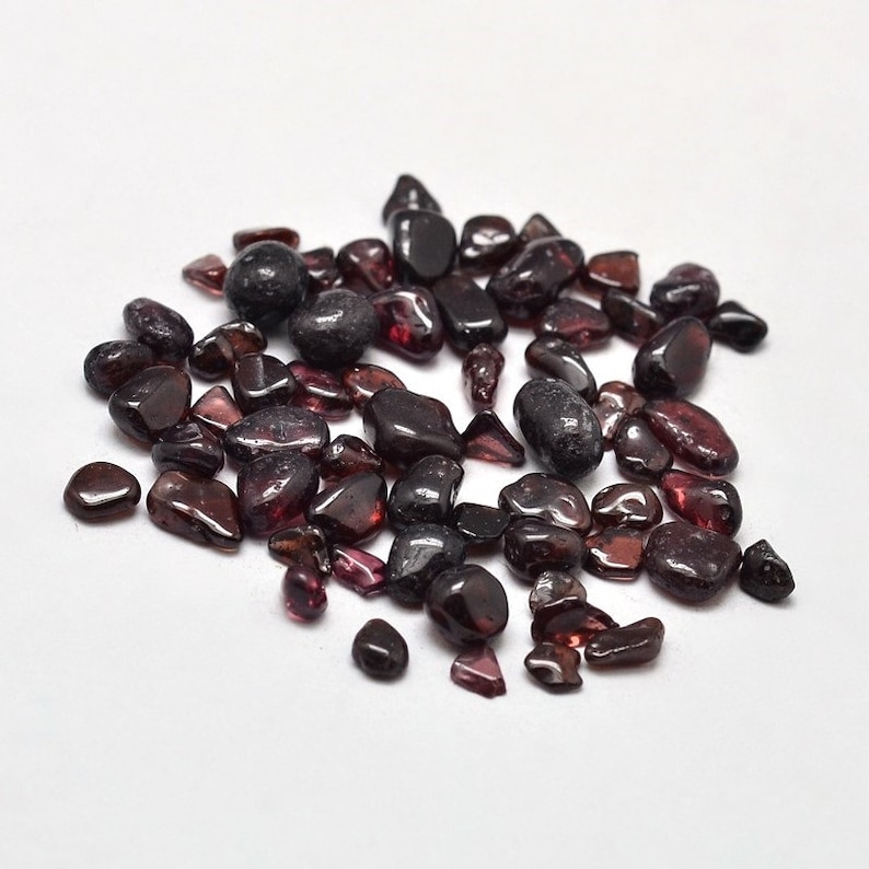 May include: A collection of polished, dark red garnet gemstones of varying sizes and shapes. The gemstones are scattered on a white surface, with some appearing translucent and others opaque. The overall appearance is smooth and reflective.