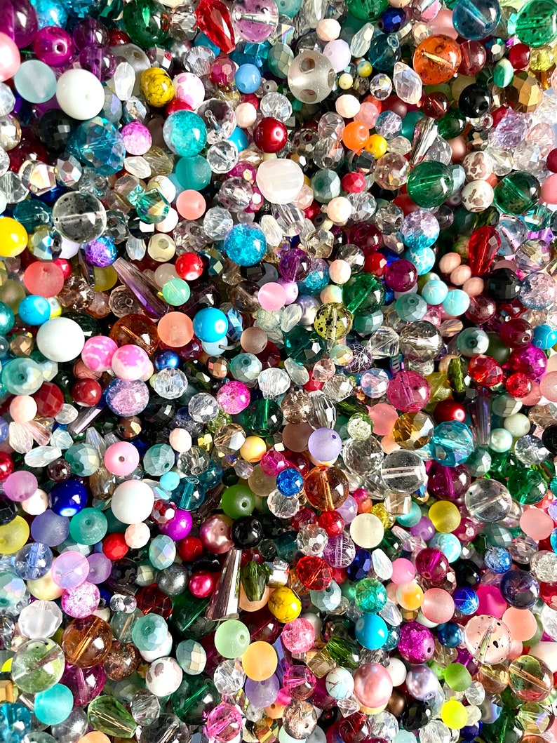 Op de afbeelding: Een grote verzameling kleurrijke glazen kralen in verschillende vormen en maten. De kralen zijn een mix van heldere, ondoorzichtige en iriserende kleuren, waaronder blauw, groen, rood, roze en paars.