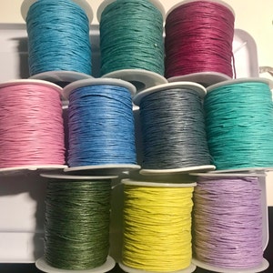 May include: Ten spools of waxed cotton cord in various colours, including teal, pink, blue, grey, green, yellow, and purple. The spools are arranged in a circle on a white surface.