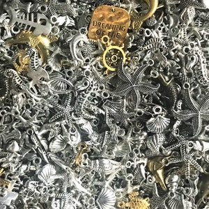 May include: A large assortment of silver and gold tone metal charms with nautical and ocean themes. The charms include seahorses, starfish, anchors, seashells, and a charm with the text "Dreaming of the Sea".